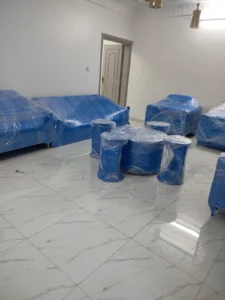 "أثاث مغلف بالبلاستيك الأزرق ومُرتب داخل غرفة بأرضية سيراميك لامعة، يظهر باب مفتوح في الخلفية."